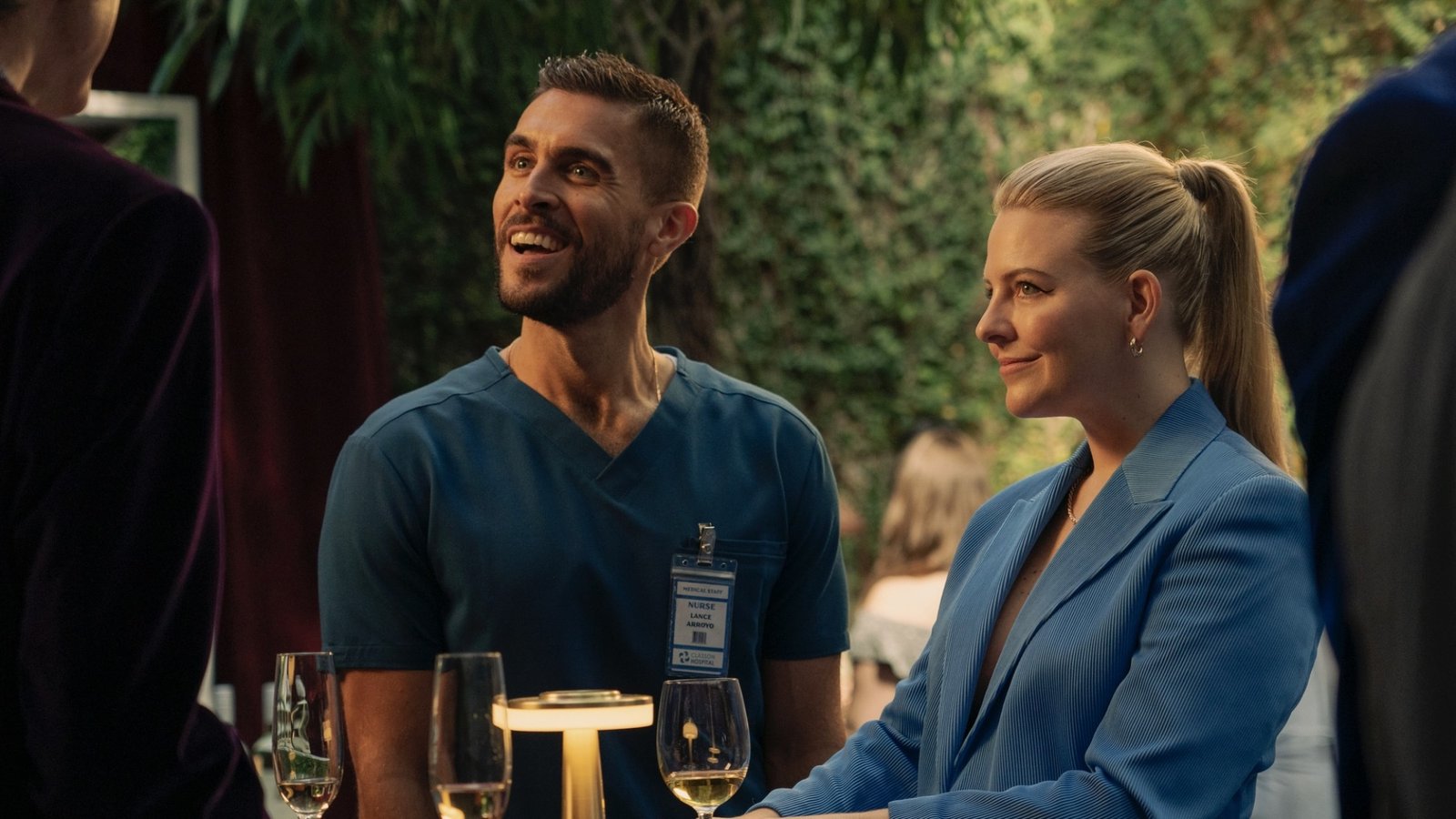 Lance in blue nurse scrubs and Brooke in a blue suit jacket at a party in “The Other Two.”