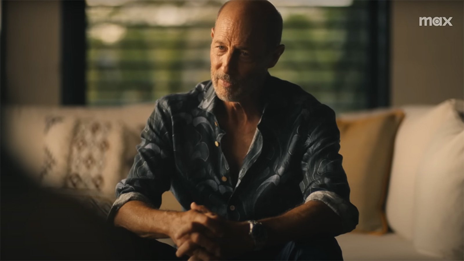 A man sits on a sofa inside, looking serious.